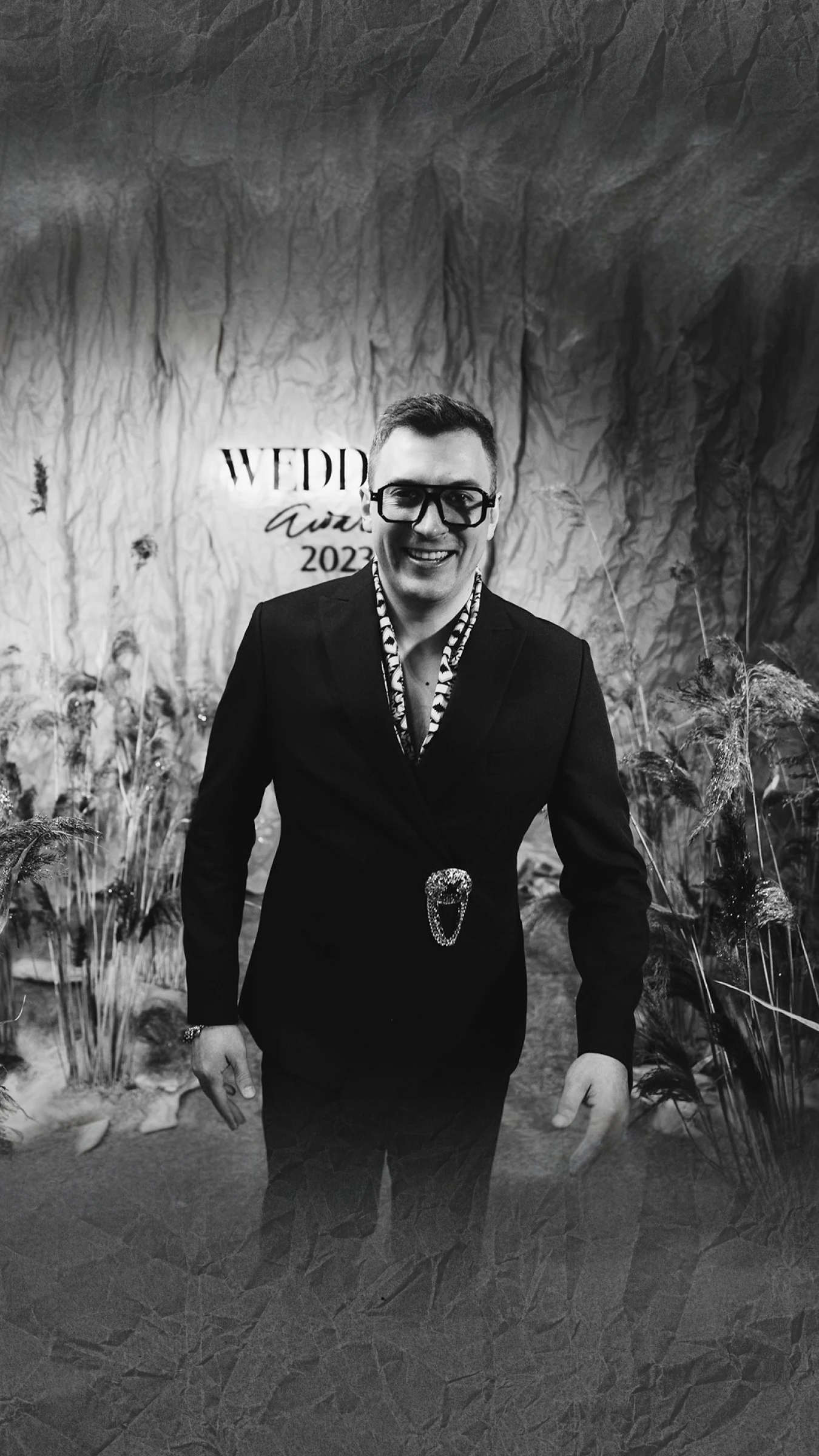 Ведущий в очках и пиджаке на фоне декораций премии Wedding Awards