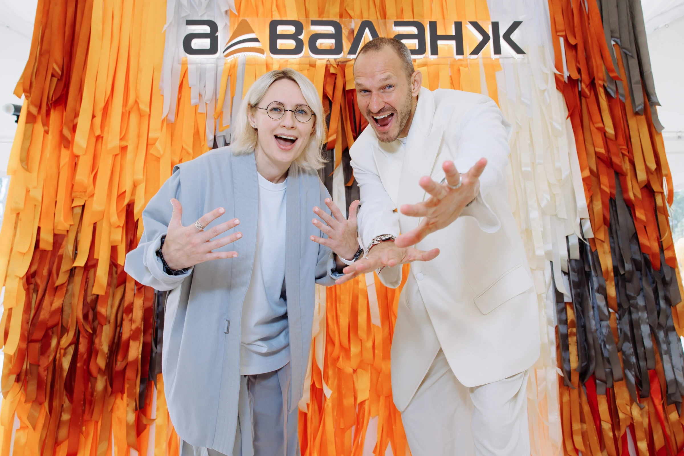 Ведущие позируют у оранжевого бахромчатого баннера с логотипом бренда