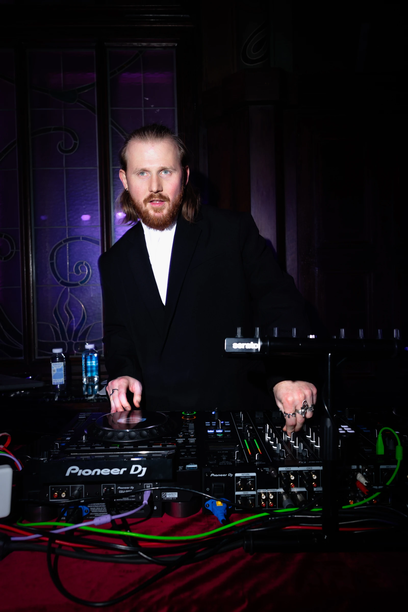Диджей в смокинге стоит за пультом Pioneer на фоне витражного окна