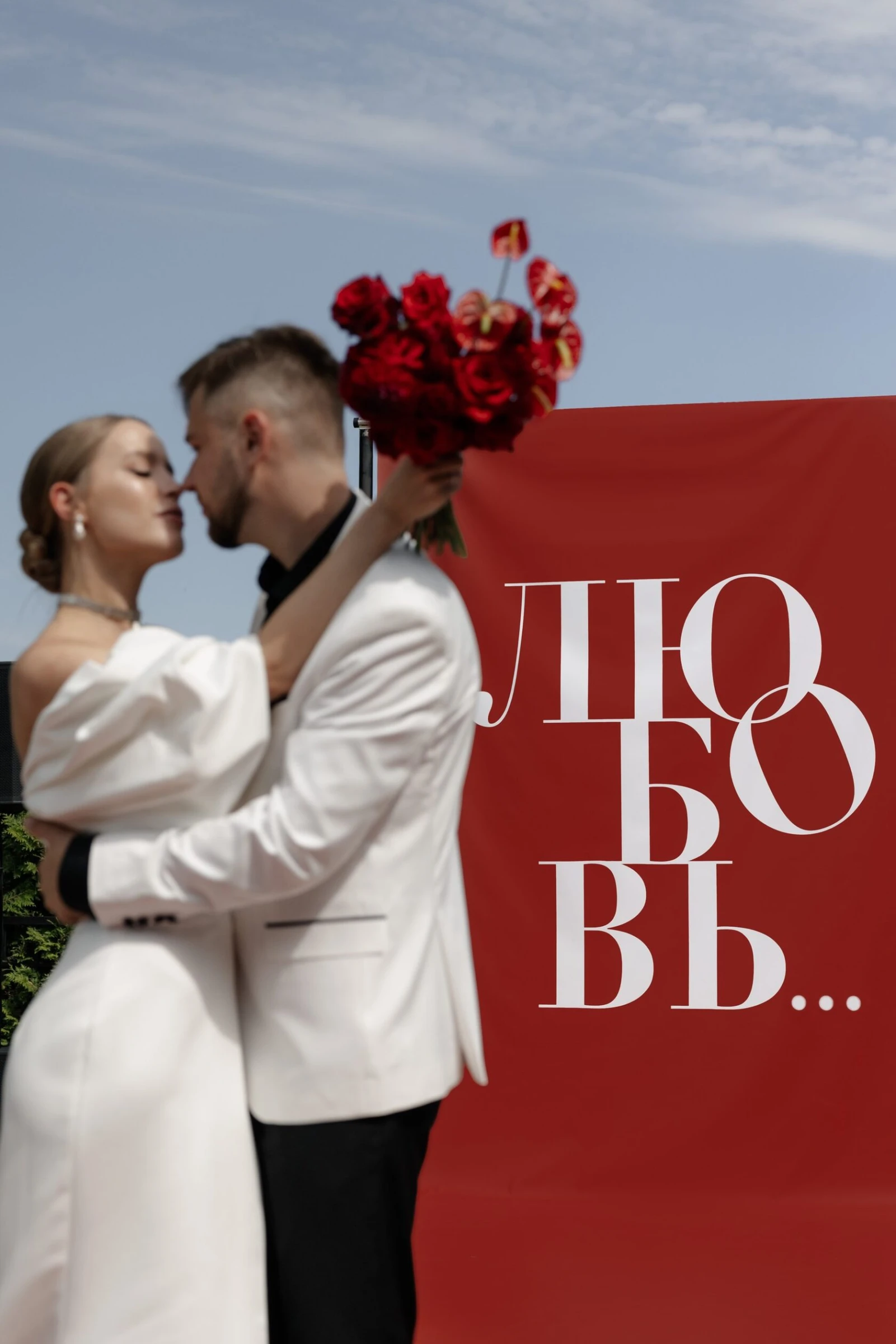 Жених и невеста целуются на фоне красного баннера держа букет алых роз