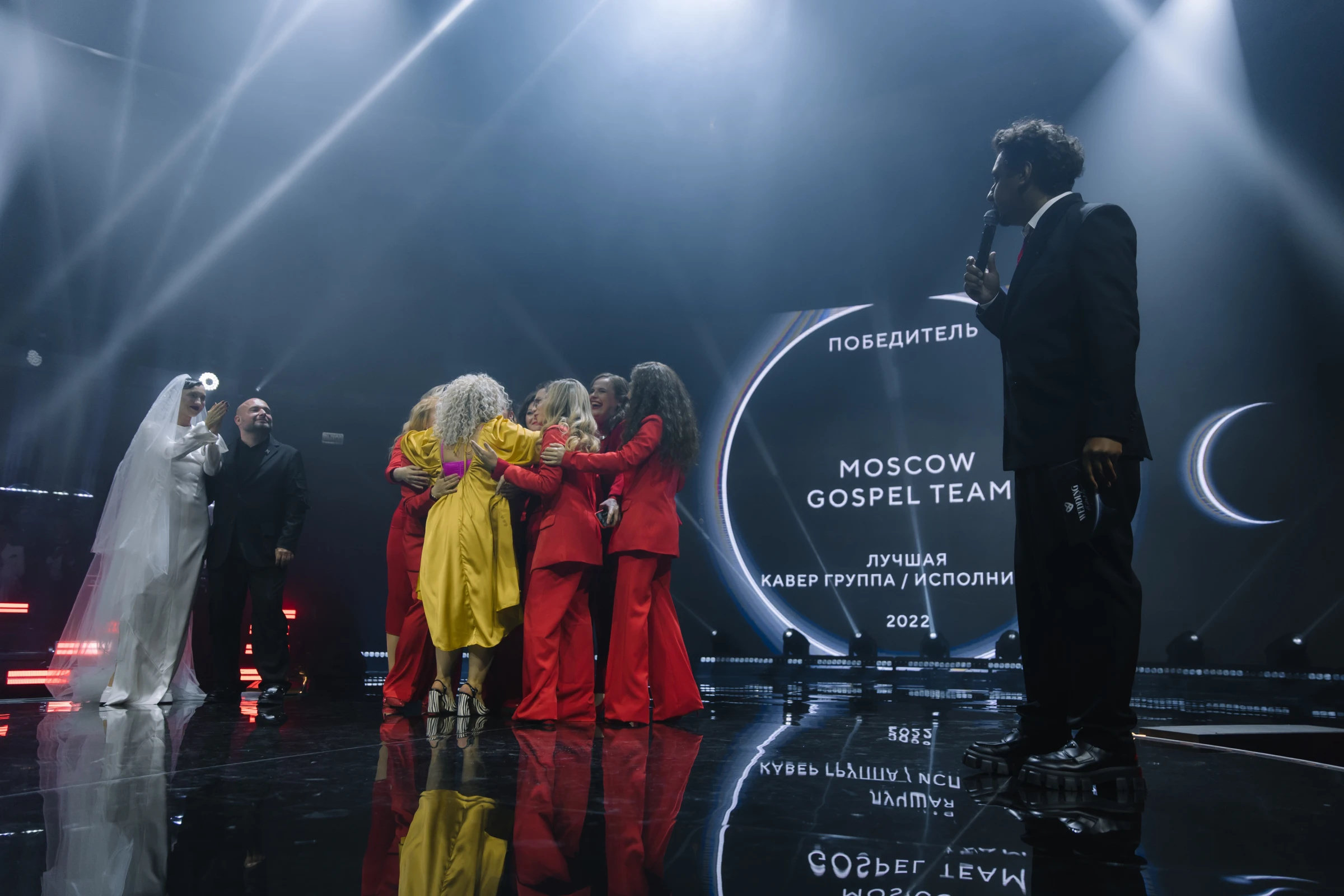 кавер-группа в красных костюмах обнимается на сцене с наградой в свете софитов