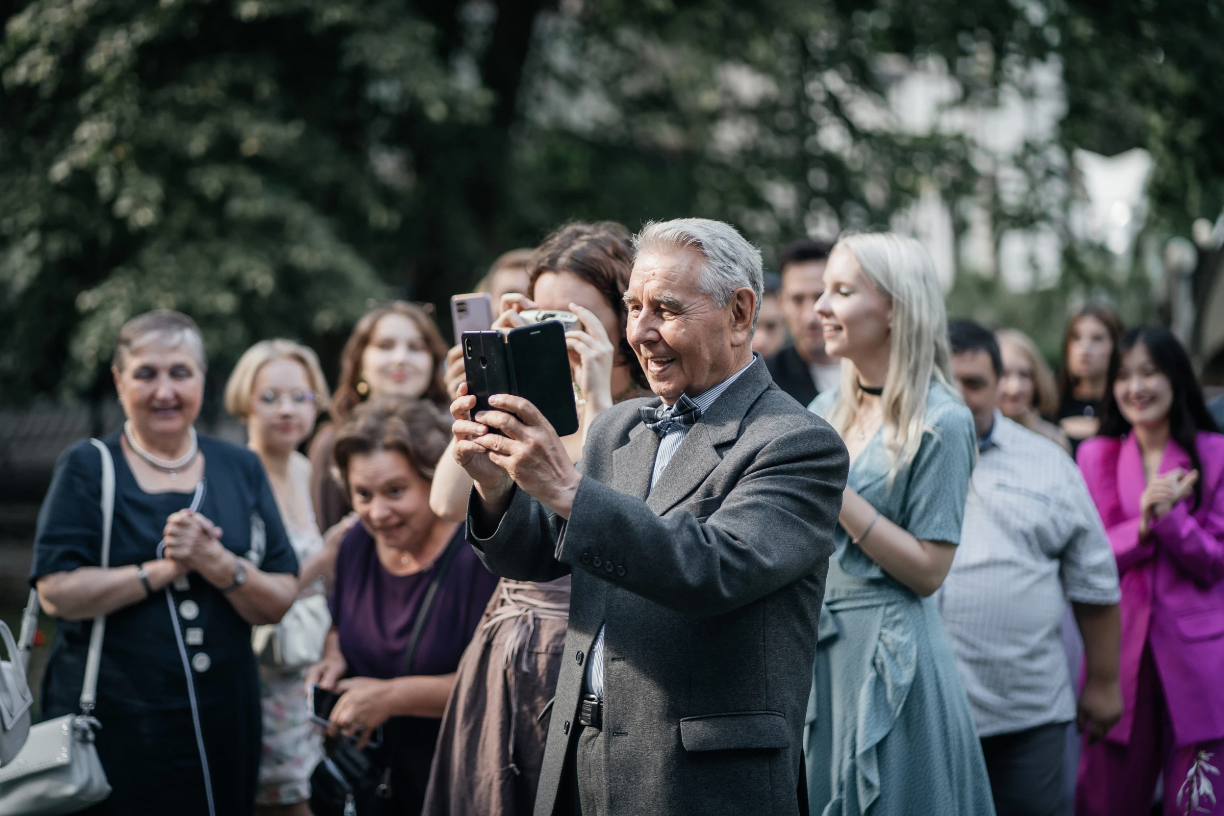 Гость в костюме фотографирует церемонию на смартфон среди зелени