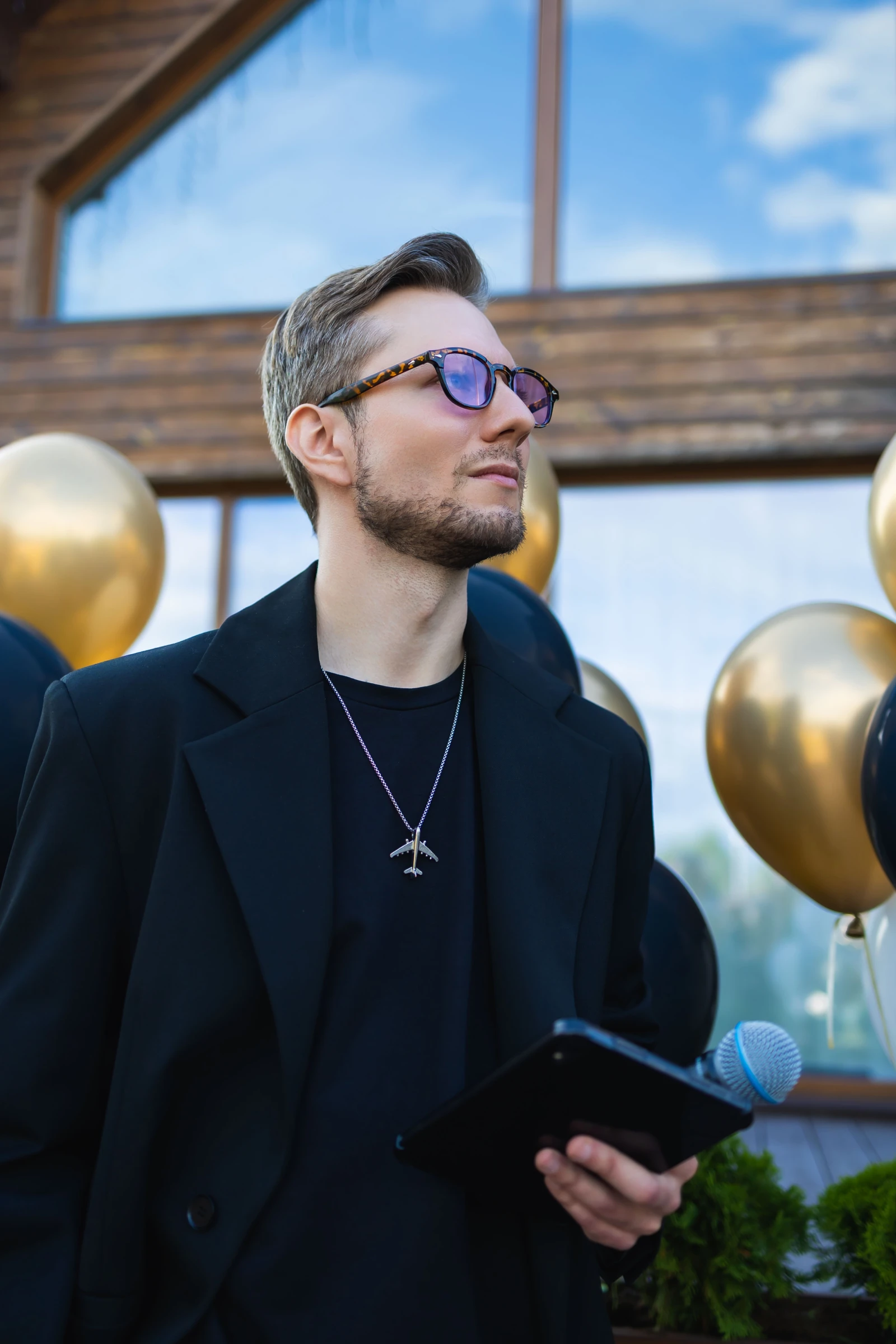 Ведущий в тёмных очках с планшетом на фоне золотых воздушных шаров