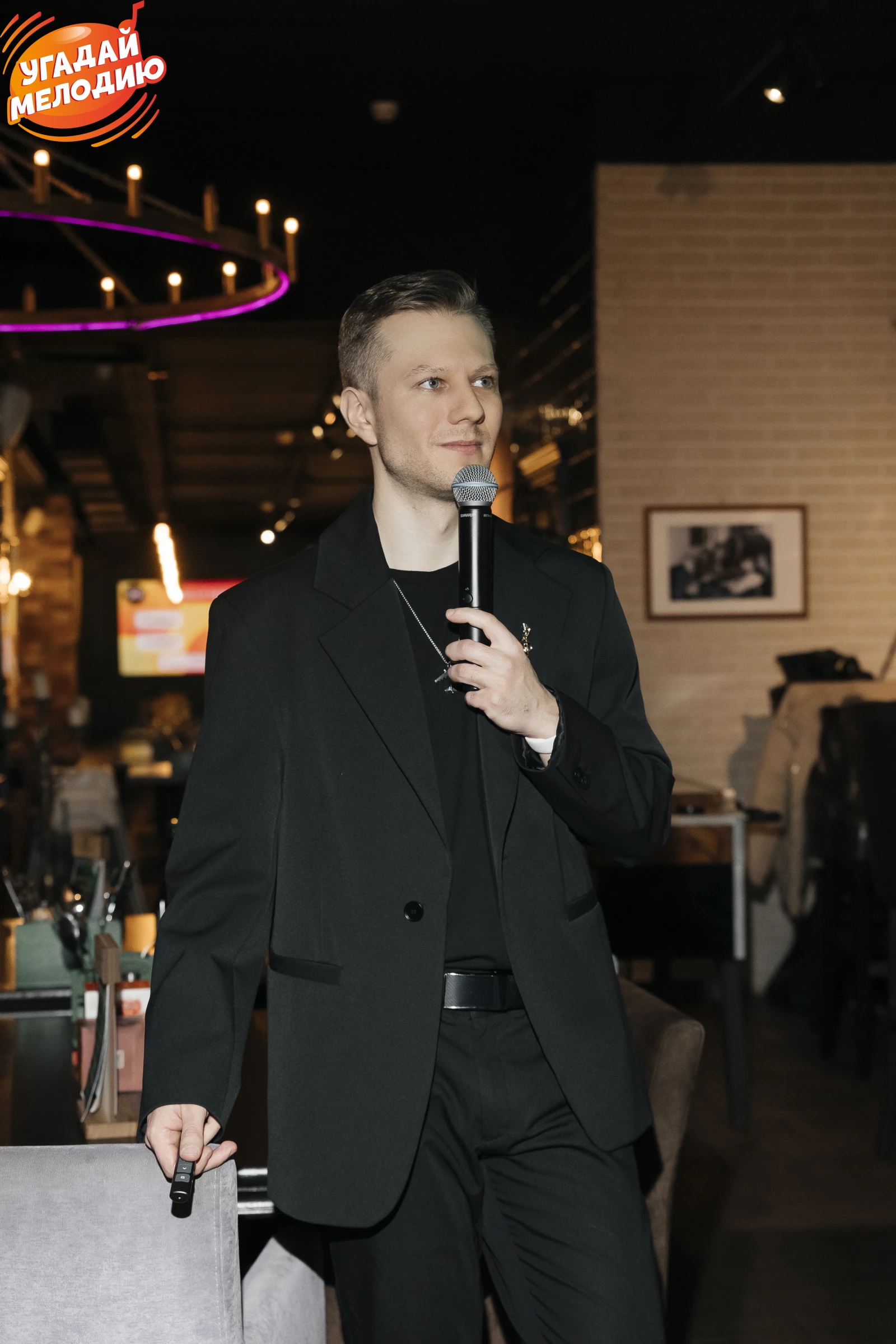 Ведущий в чёрном костюме с микрофоном на фоне барной стойки ресторана