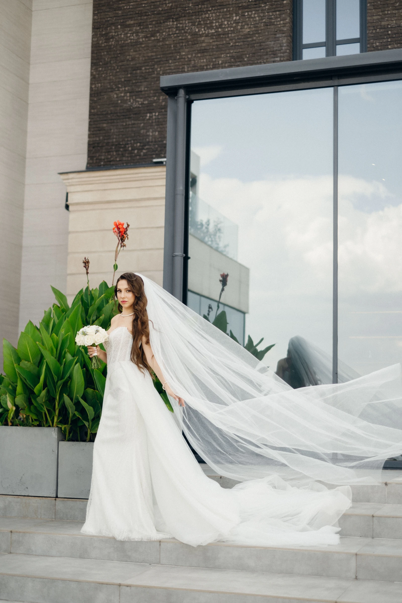 невеста с длинной фатой и букетом роз у панорамного окна площадки