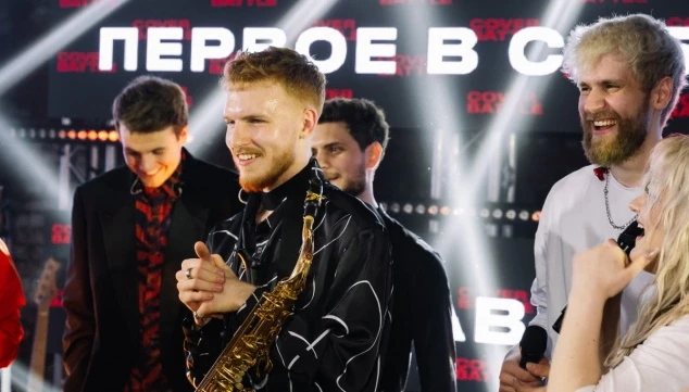 саксофонист кавер-группы в чёрном плаще стоит на фоне светового экрана