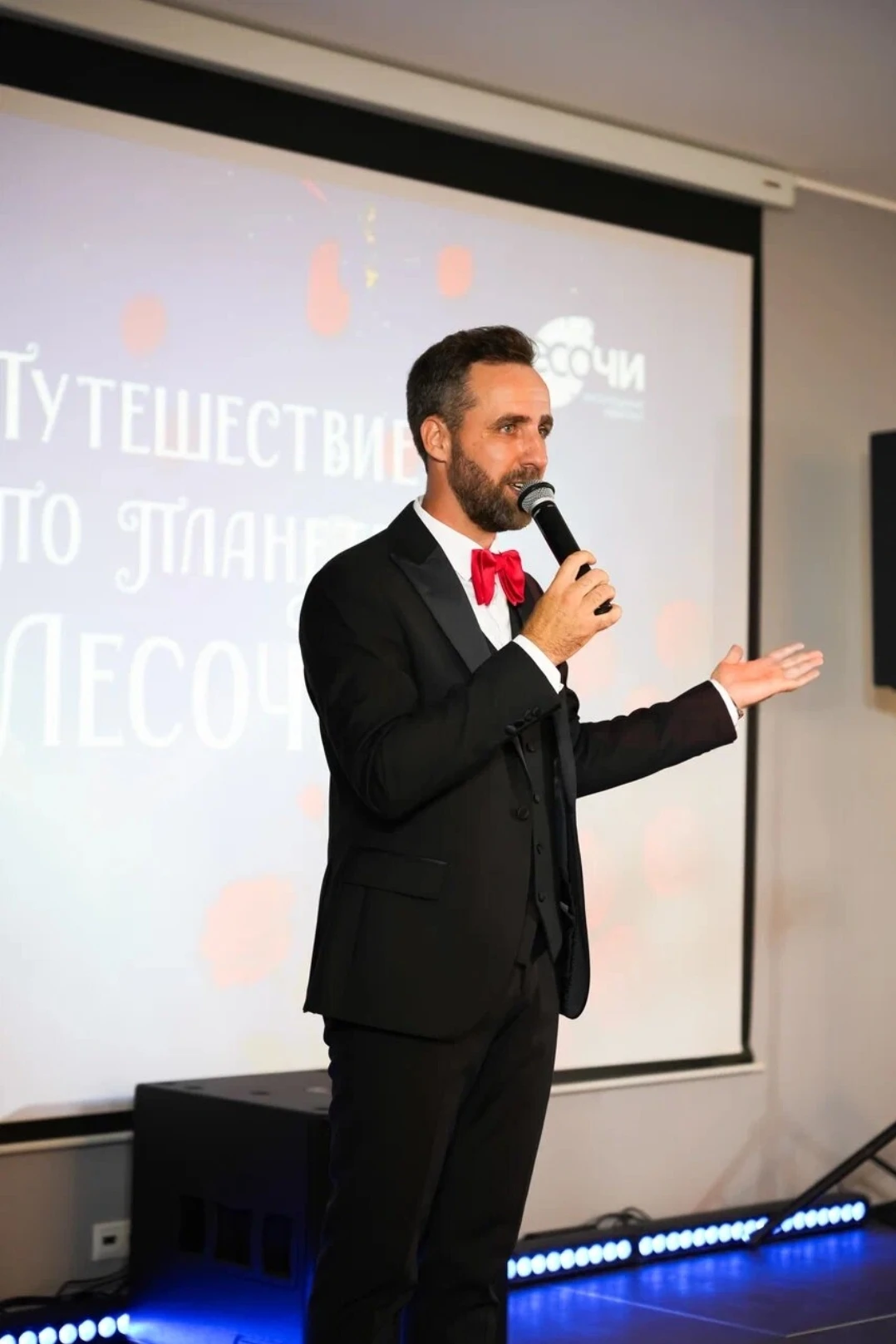 ведущий с микрофоном произносит речь на фоне экрана