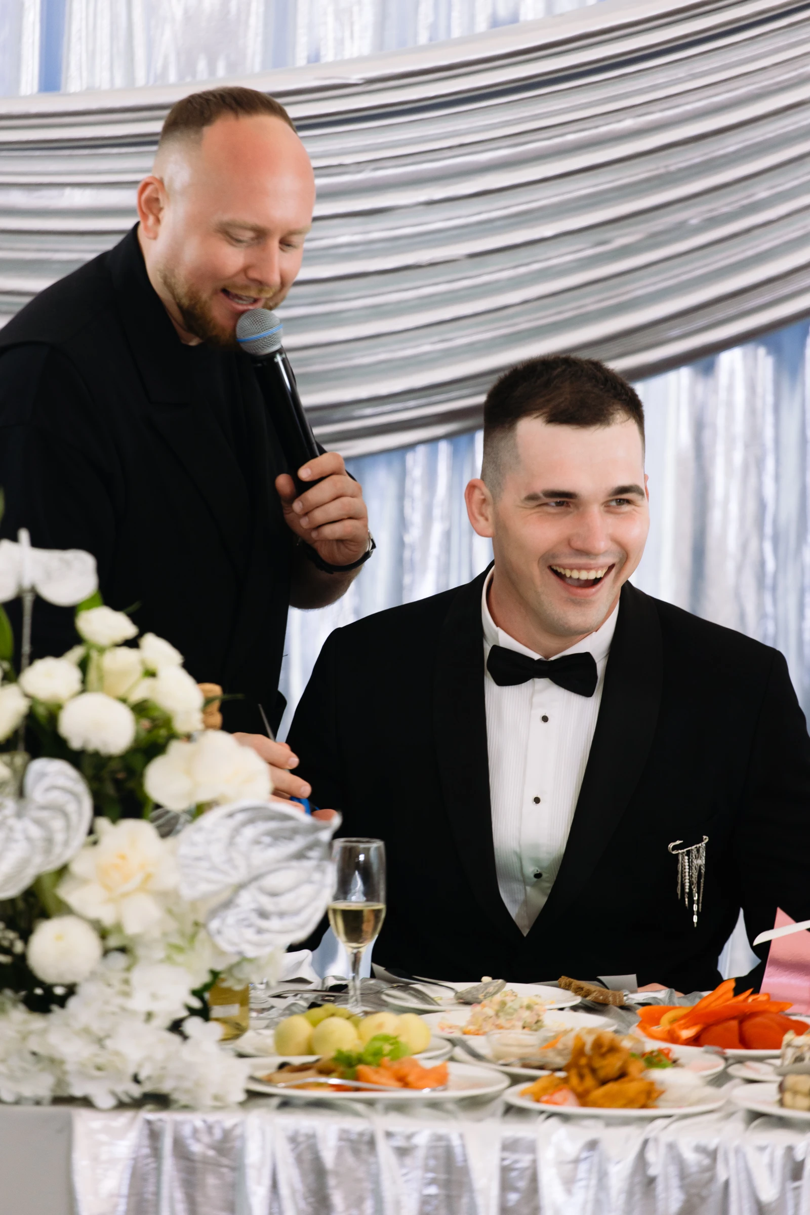 ведущий с микрофоном рядом с улыбающимся женихом за столом