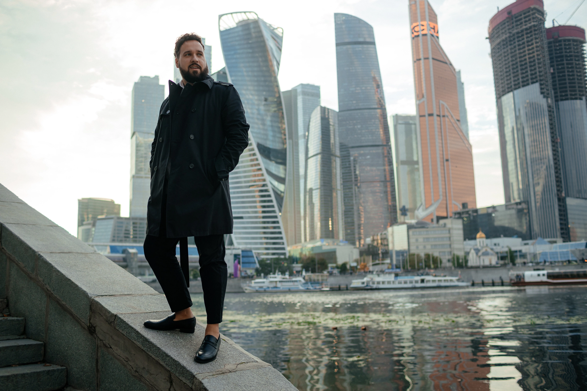 мужчина в пальто стоит у набережной на фоне Москва-Сити