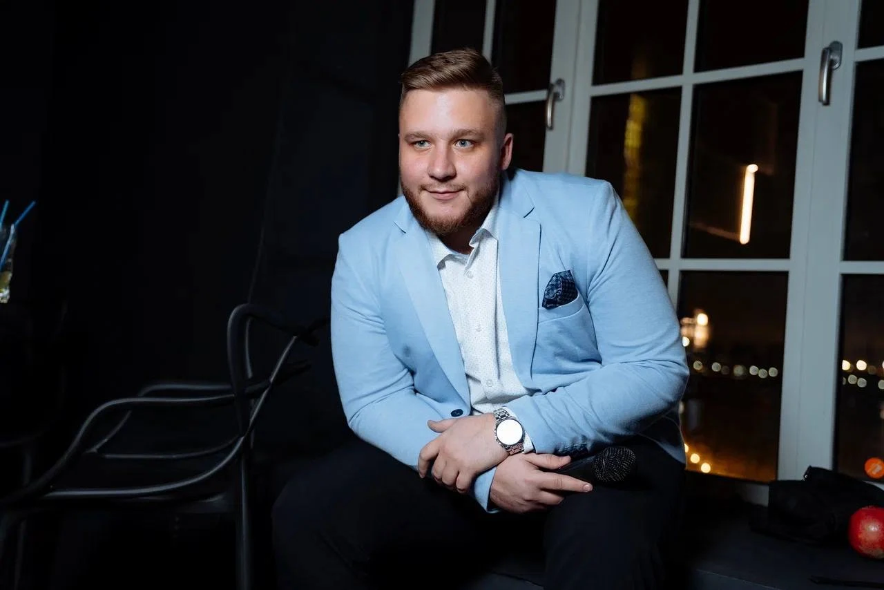 ведущий в голубом пиджаке на фоне вечерних окон банкетного зала