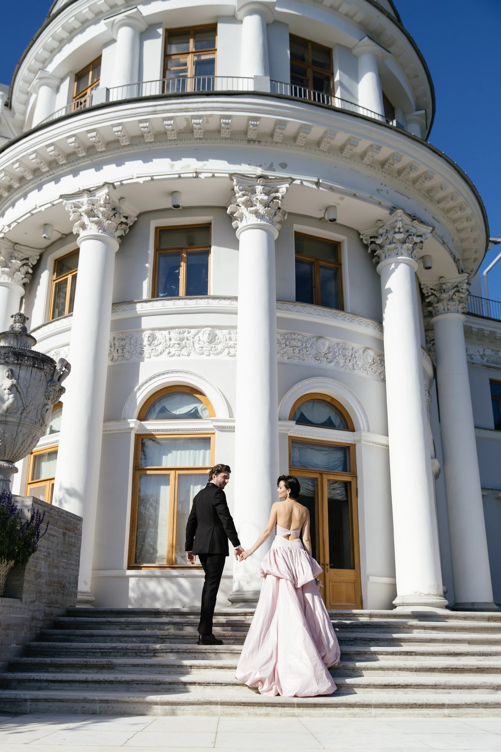 пара держится за руки на ступенях у колонн особняка днём