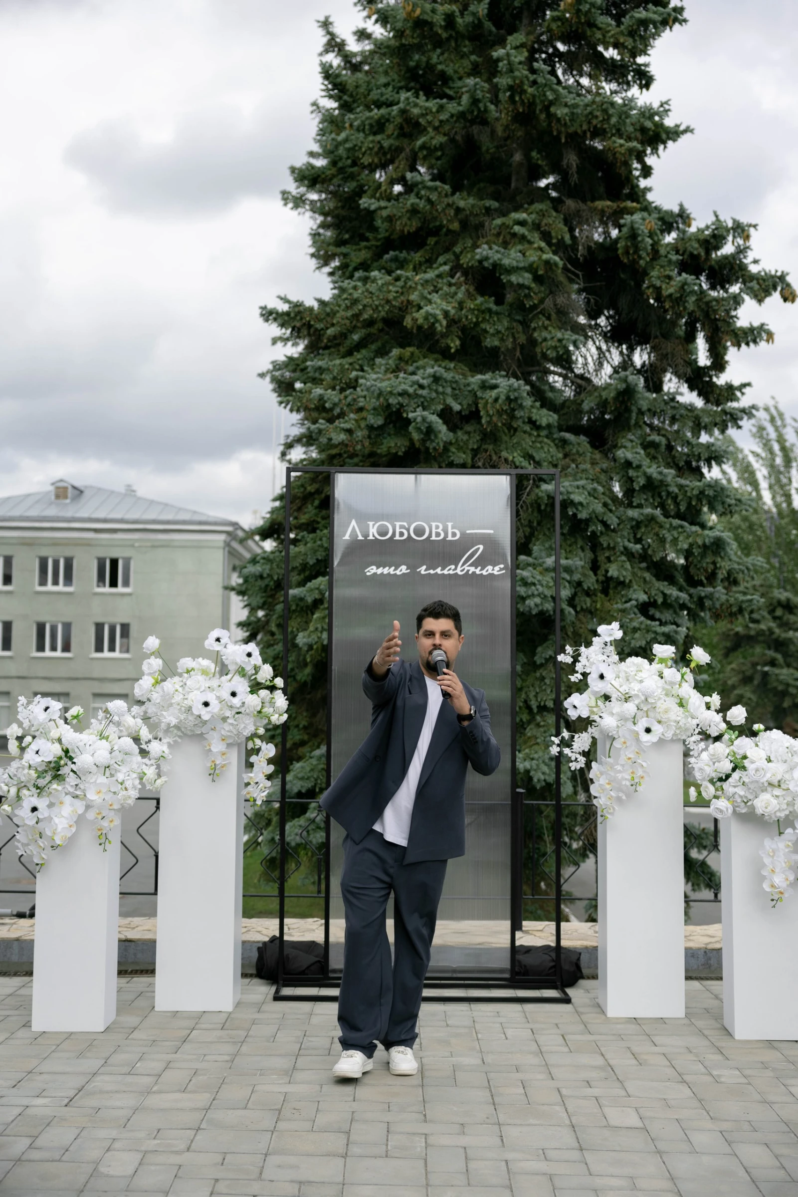 ведущий выступает с микрофоном у баннера с надписью среди белых цветочных колонн