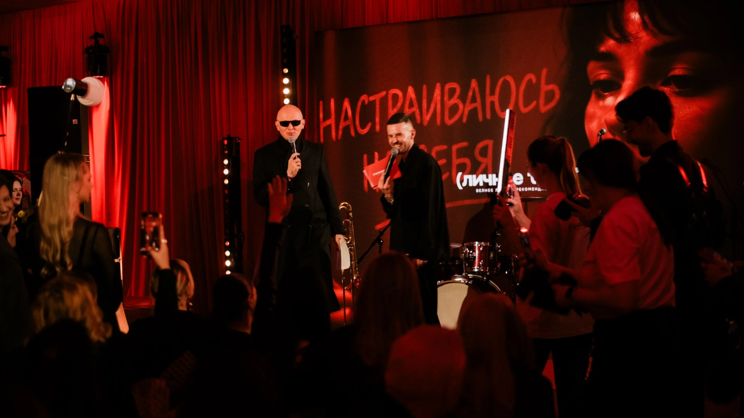 Двое ведущих с микрофонами на сцене банкета на фоне экрана с надписью