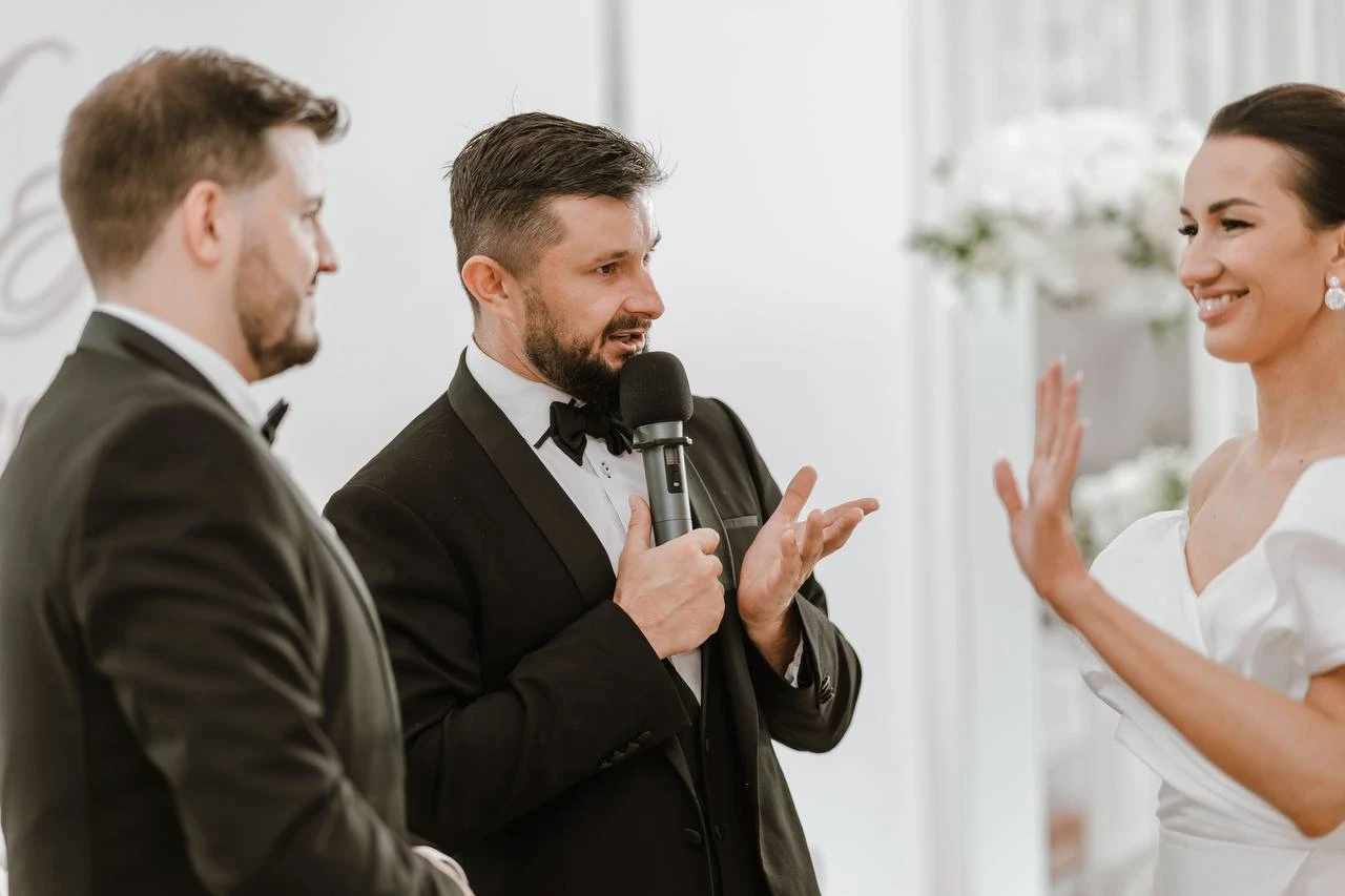 ведущий с микрофоном общается с женихом и невестой у цветочного декора
