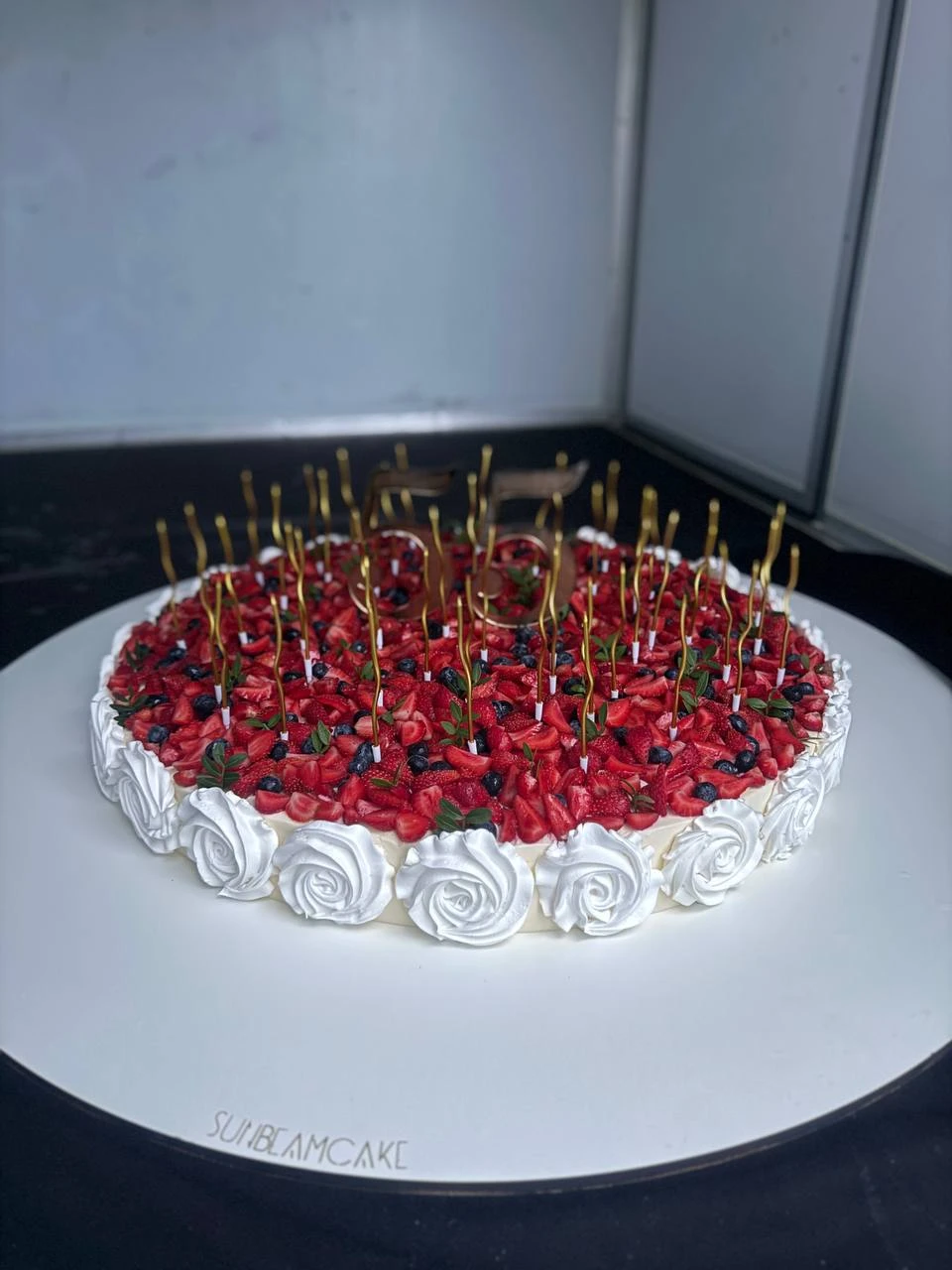 торт с кремовыми розами клубникой черникой и золотыми свечами на белом постаменте