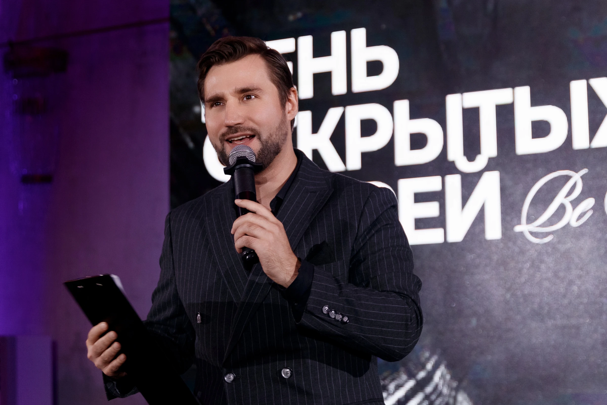 ведущий с микрофоном и папкой выступает на сцене мероприятия
