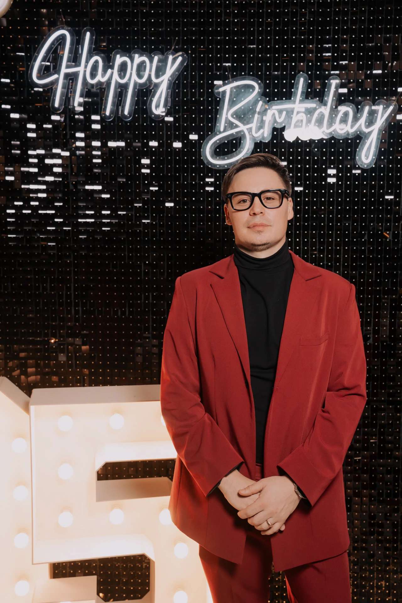 ведущий в красном костюме позирует на фоне неоновой вывески Happy Birthday