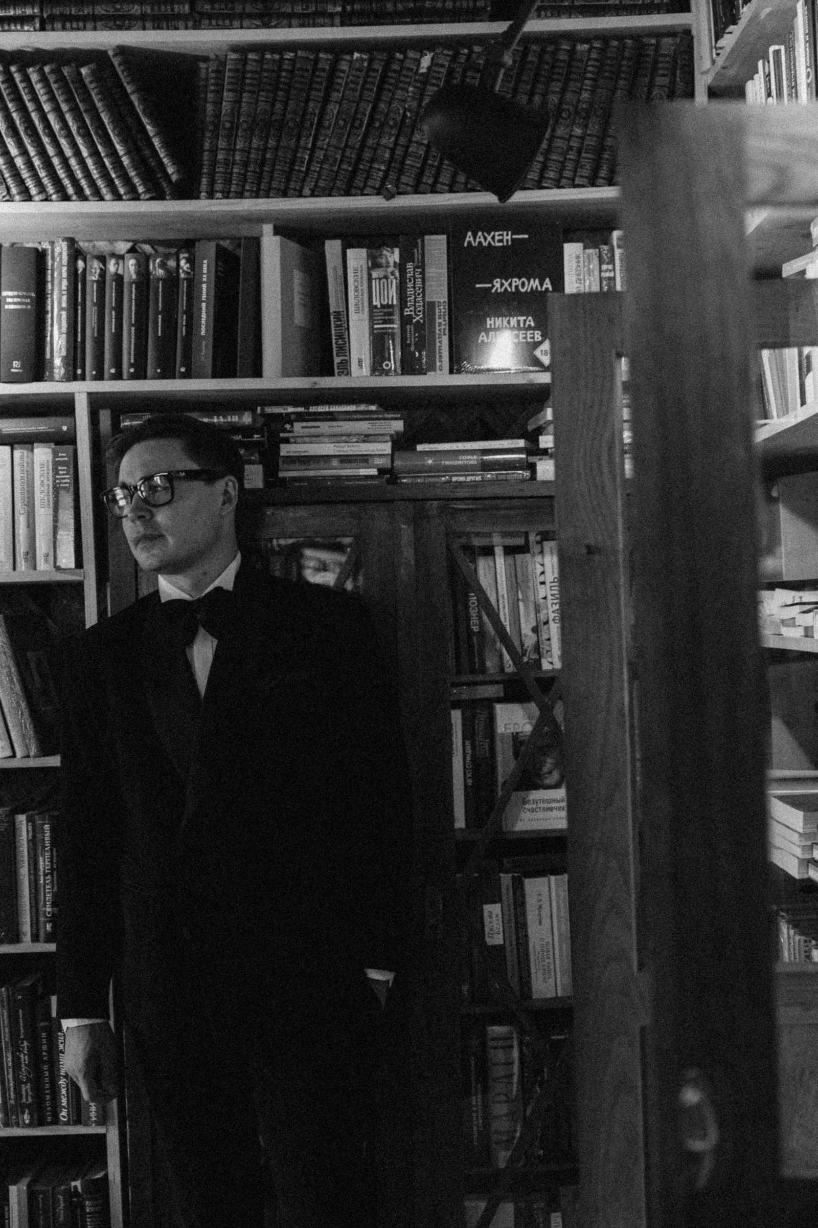 ведущий в чёрном смокинге и очках стоит у книжных стеллажей библиотеки
