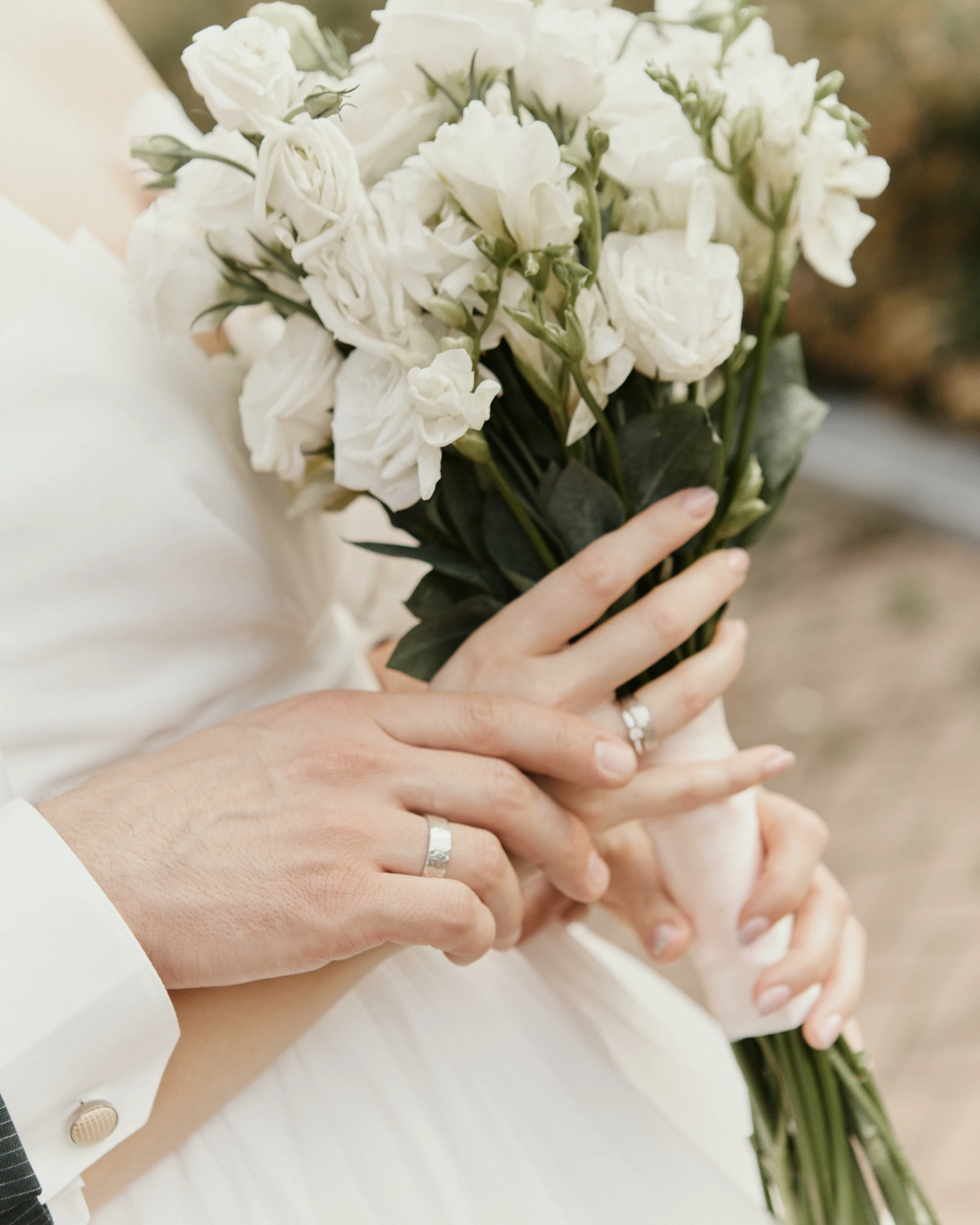 переплетённые руки жениха и невесты с обручальными кольцами возле букета белых роз