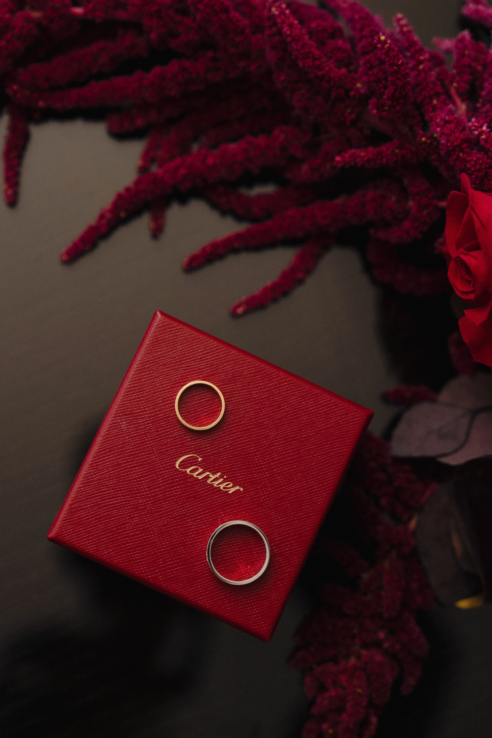 обручальные кольца на красной коробке Cartier среди бордовых цветов