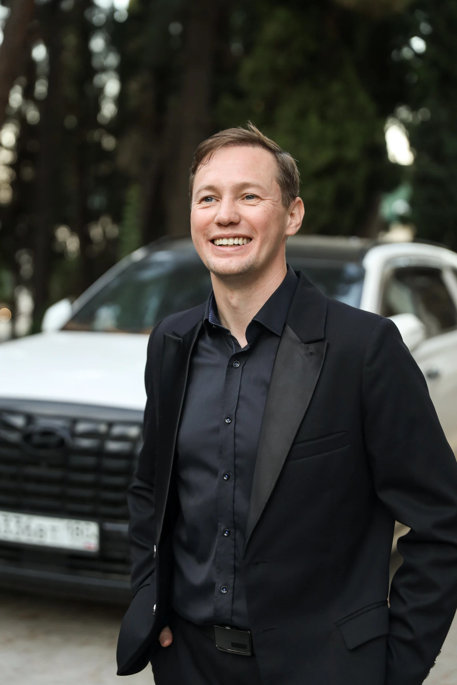 улыбающийся ведущий свадьбы в чёрном костюме у автомобилей